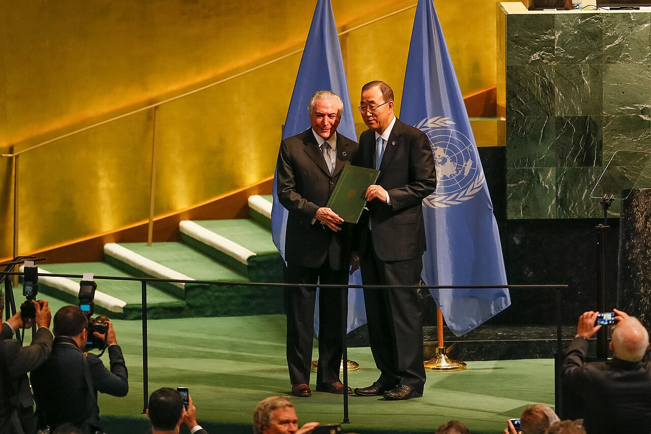 Two men hold a green folder before UN flags