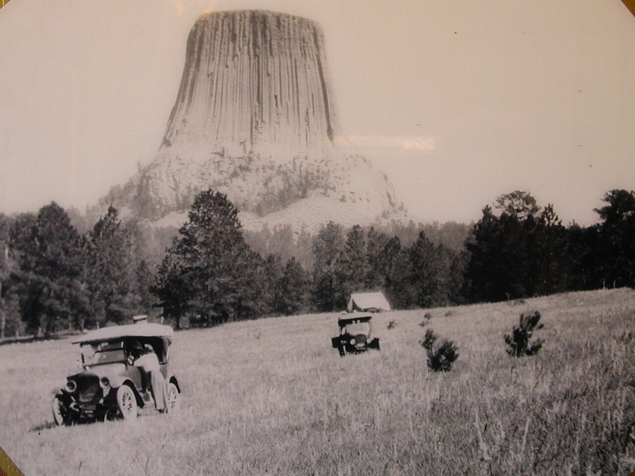 Early automobiles stand in the meadow below the tower