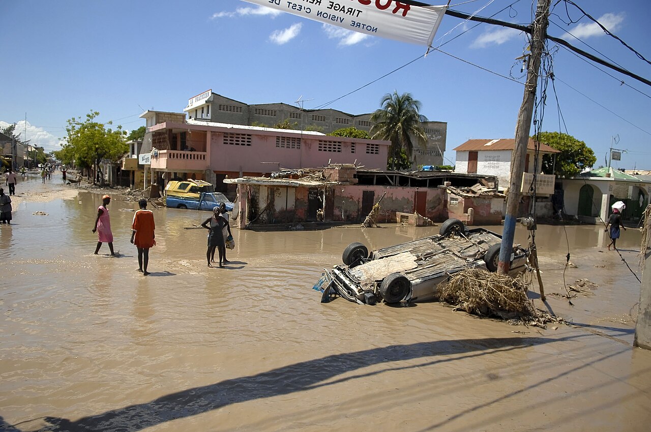 Residents walk through flooded streets where a car lies overturned